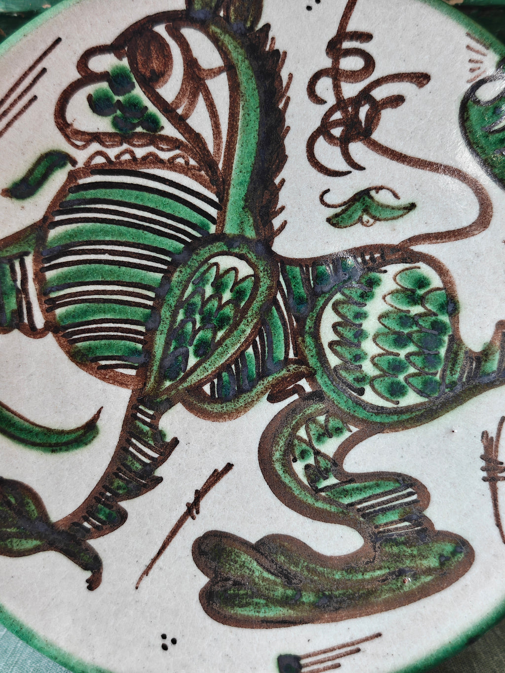 Close up of a round ceramic plate with a hand-painted green and brown dragon design on a white background.