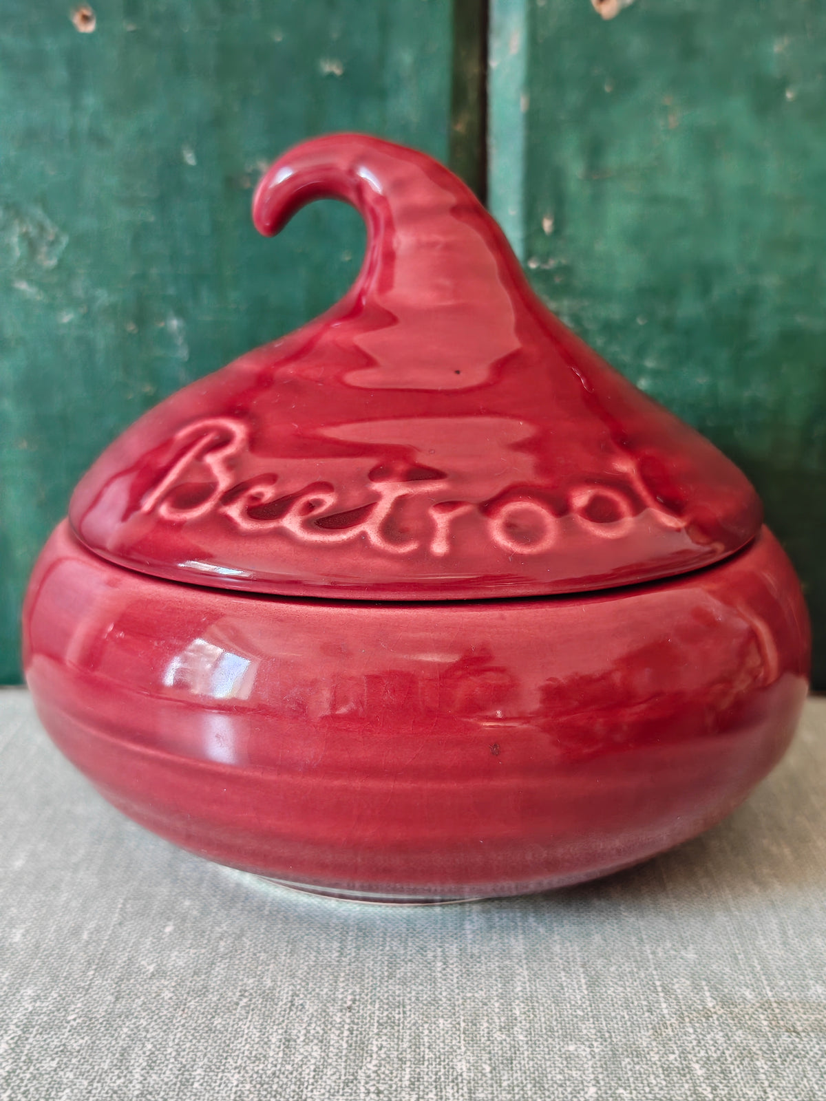 A glossy red ceramic lidded jar with a sculpted lid resembling a beetroot and embossed text.