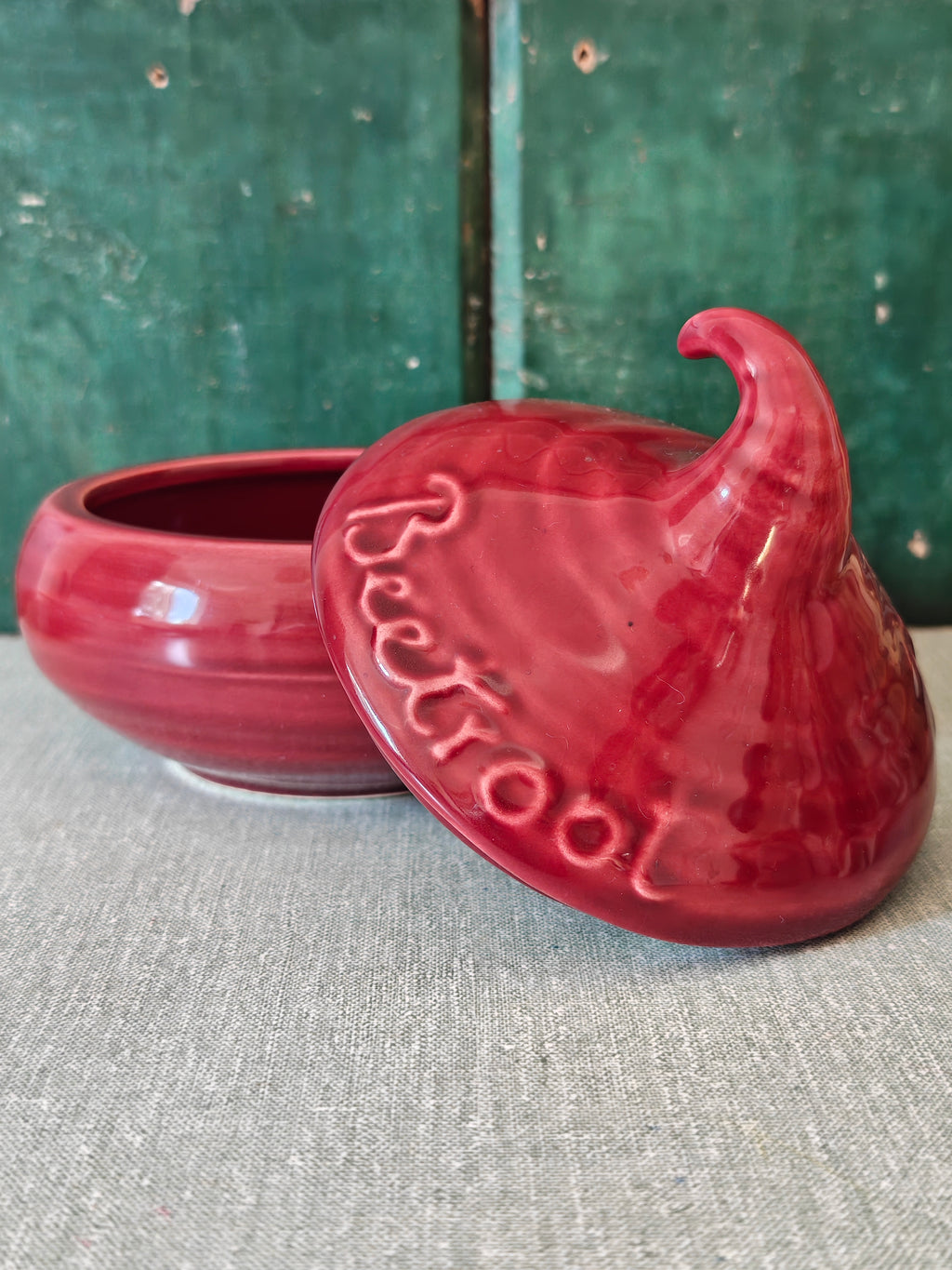 A glossy red ceramic jester tin with its lid leaning against the base. The lid features embossed text reading 'Beetroot'.