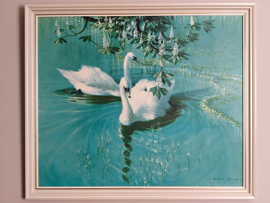 A framed painting of two white swans swimming in a turquoise pond under hanging white flowers, set in a white frame.