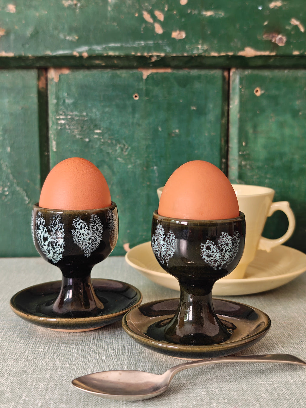 Two dark green ceramic egg cups with white floral patterns and attached saucers. 