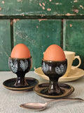 Two dark green ceramic egg cups with white floral patterns and attached saucers. 
