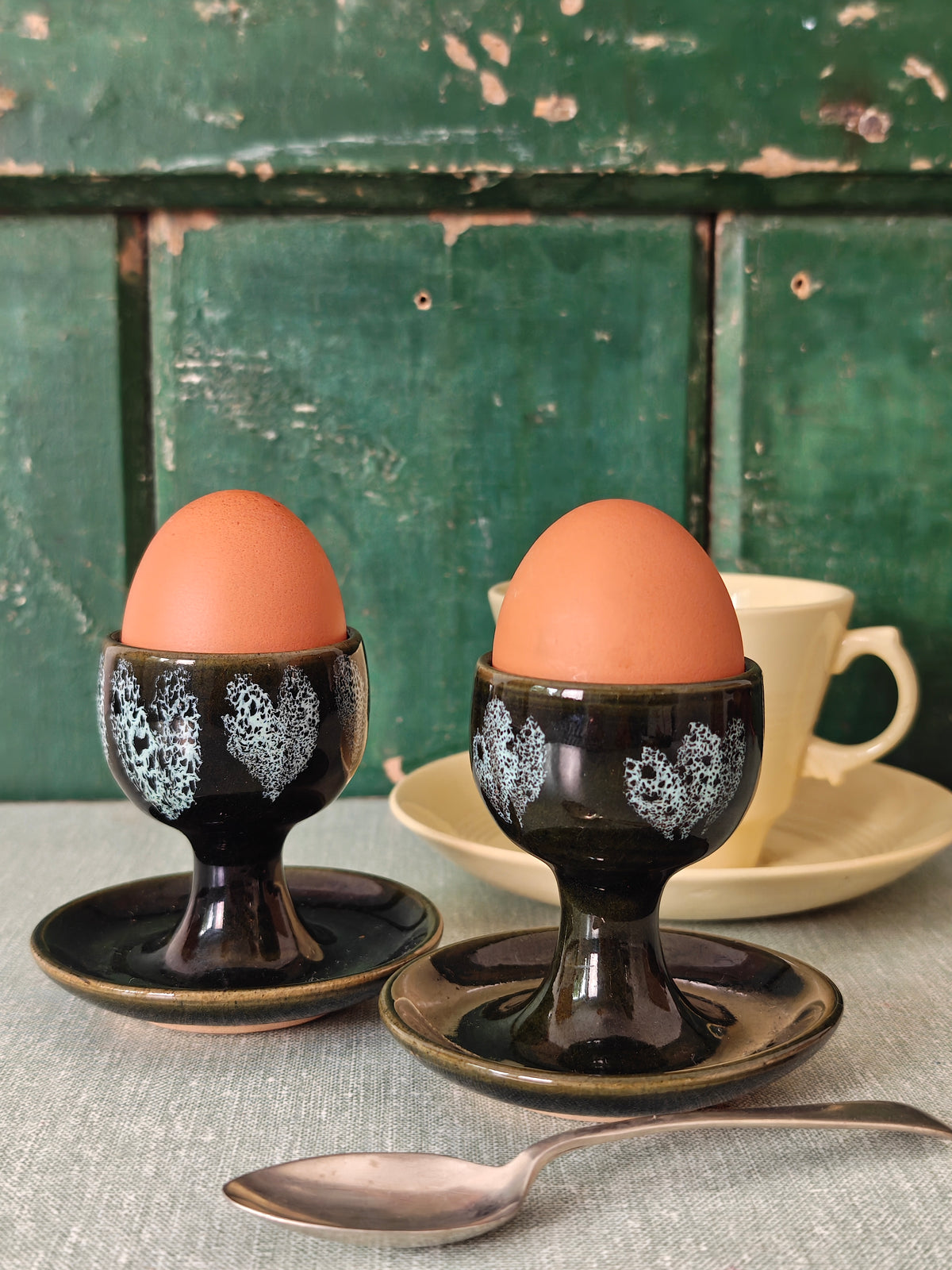 Two dark green ceramic egg cups with white floral patterns and attached saucers. 