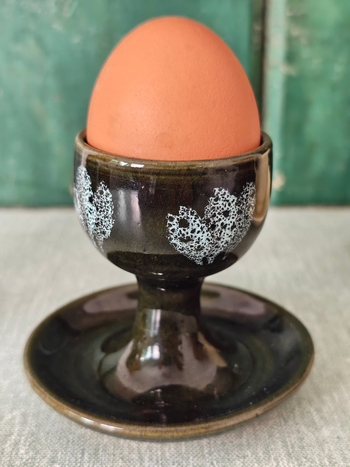 A dark green ceramic egg cup with white floral patterns and attached saucer. 