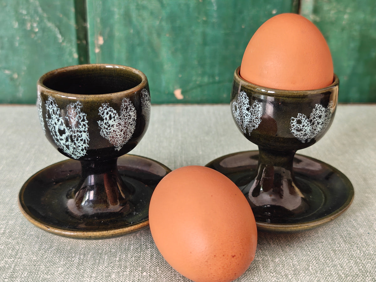 Two dark green ceramic egg cups with white floral patterns and attached saucers. 