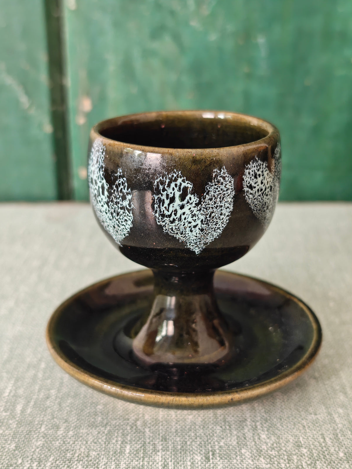 A dark green ceramic egg cup with white heart pattern and attached saucer. 