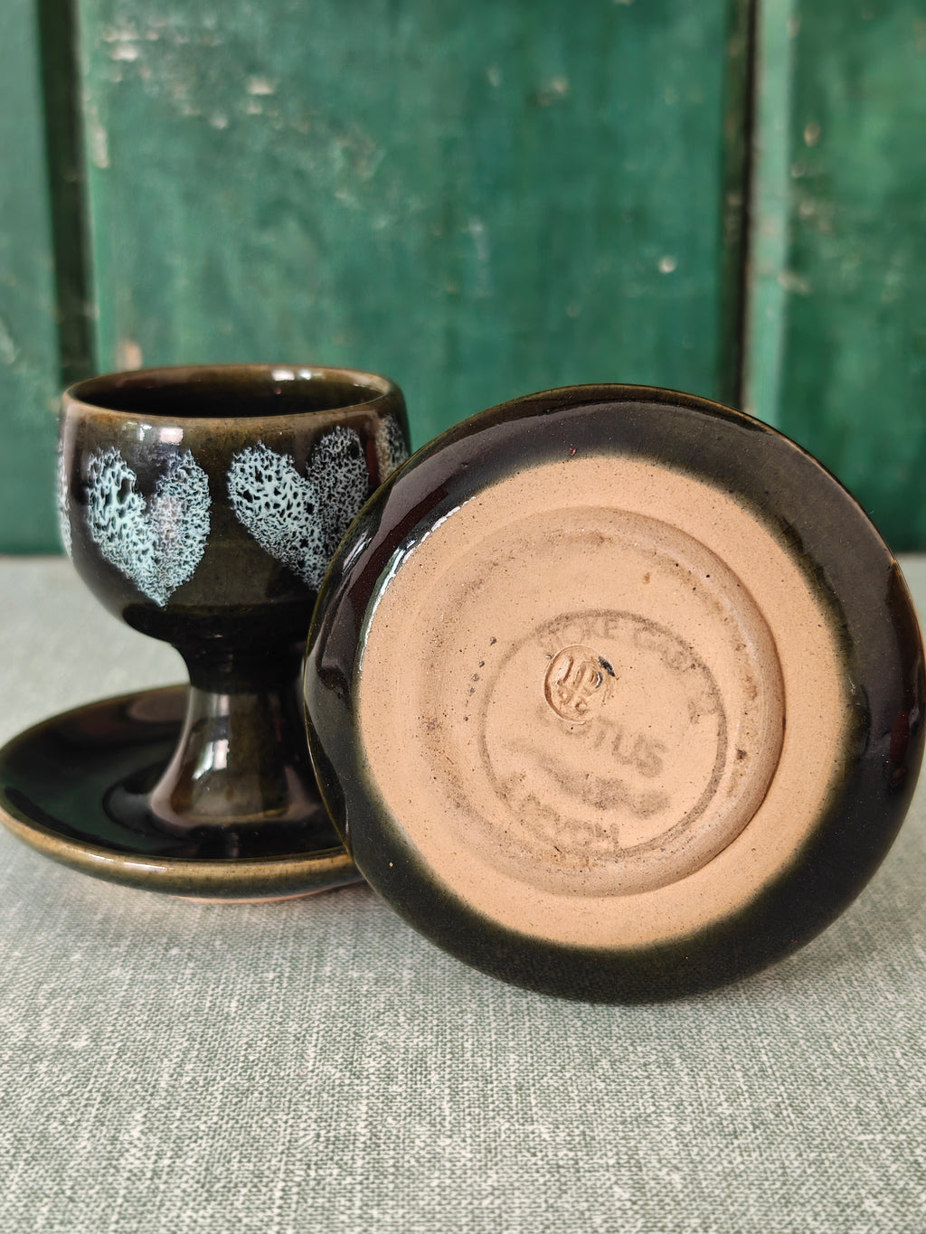 Two dark green ceramic egg cups with white floral patterns and attached saucers.  One egg cup is tipped on its side to show the makers mark underneath.