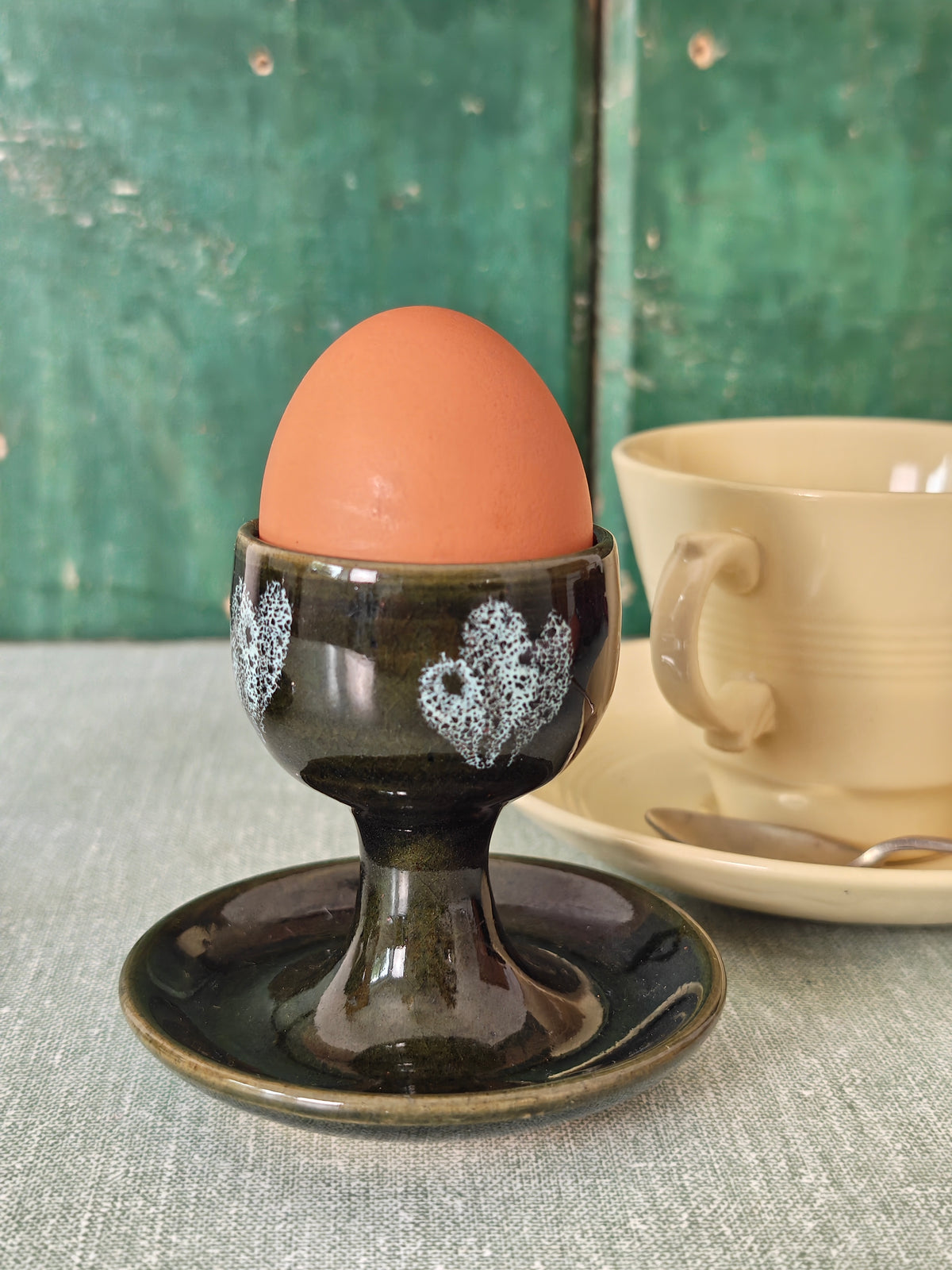 A dark green ceramic egg cups with white floral patterns and attached saucer. 