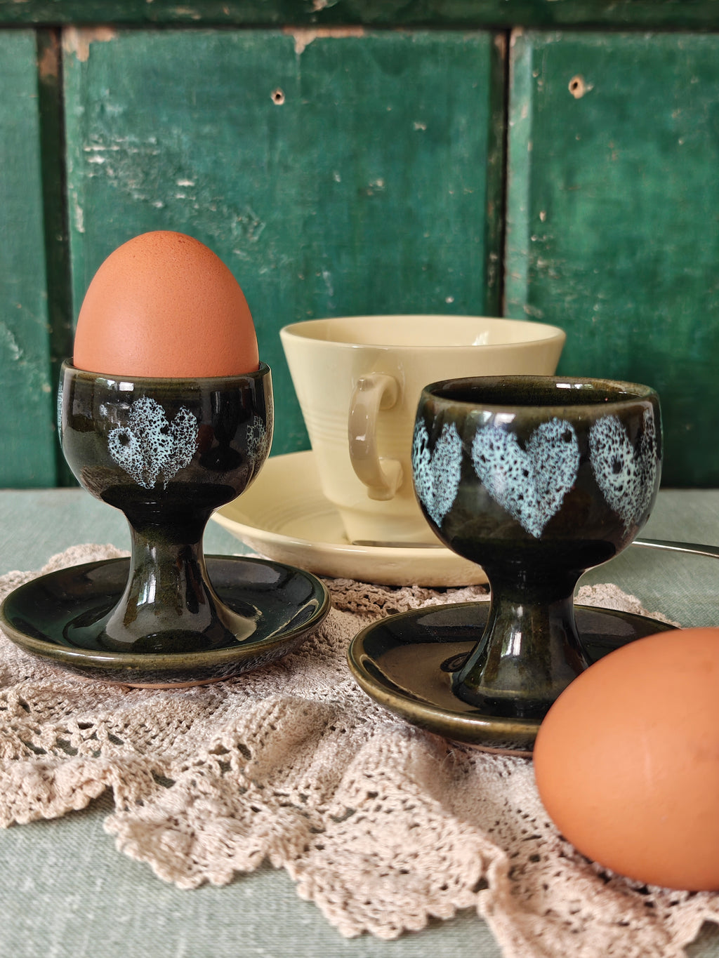 Two dark green ceramic egg cups with white floral patterns and attached saucers. 
