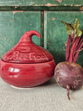 A glossy red ceramic lidded jar with a sculpted lid resembling a beetroot and embossed text.