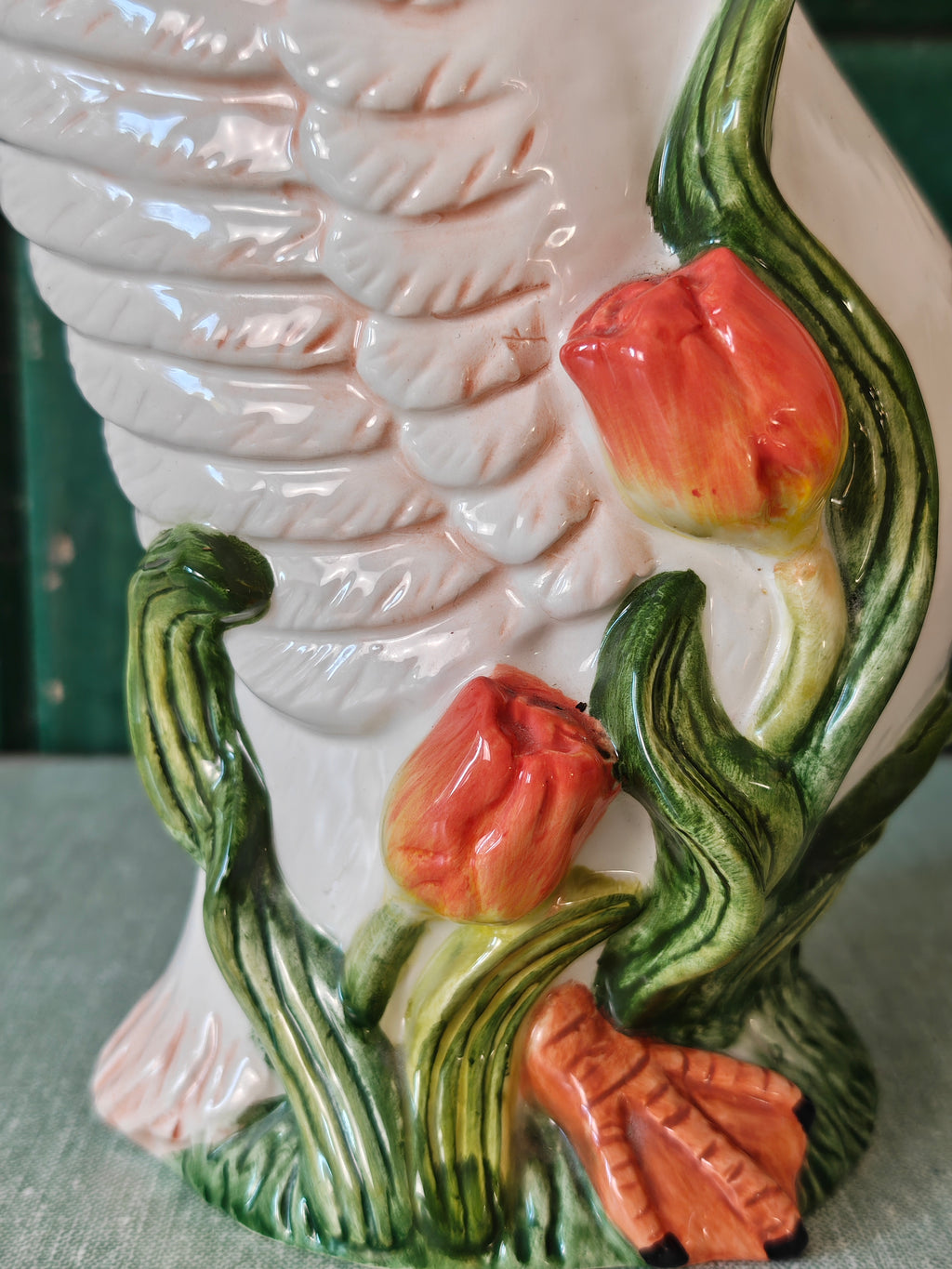 Close-up of a ceramic bird with detailed feather texture, green leafy stems, and three glossy red-orange tulip flowers.