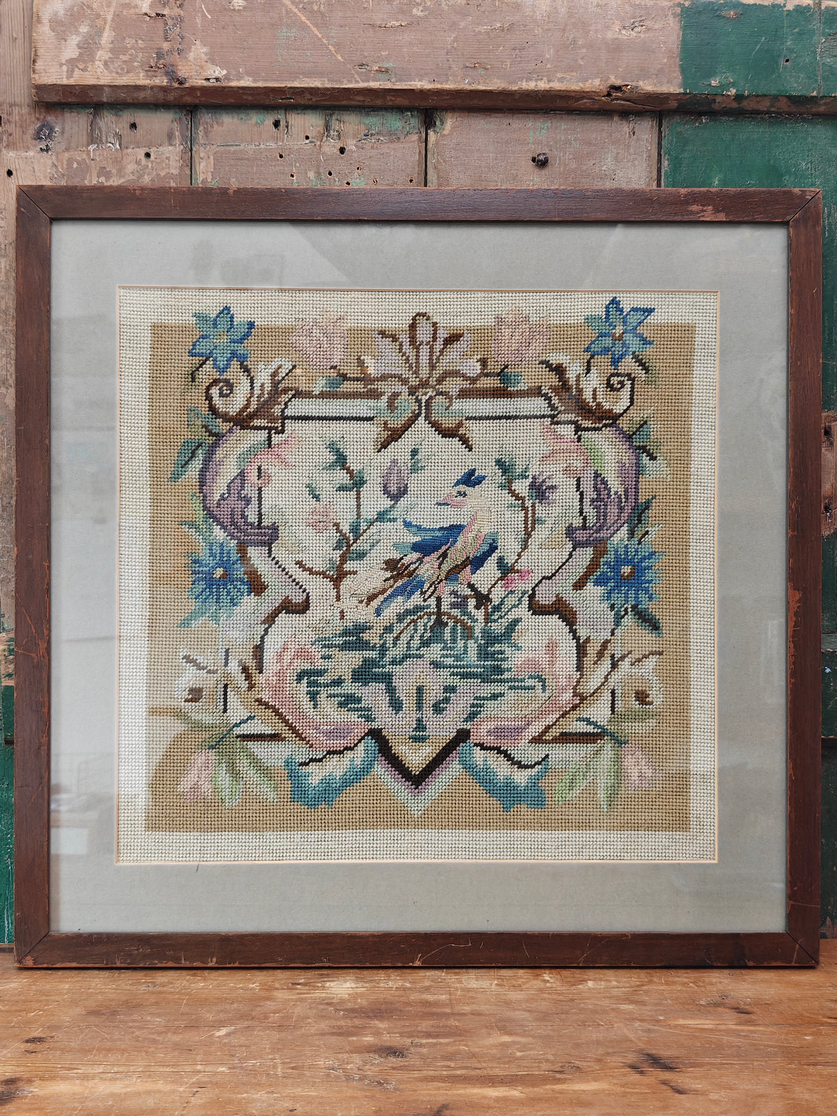 A framed needlepoint artwork featuring a bird and floral scroll design in muted blue, pink, and beige tones.