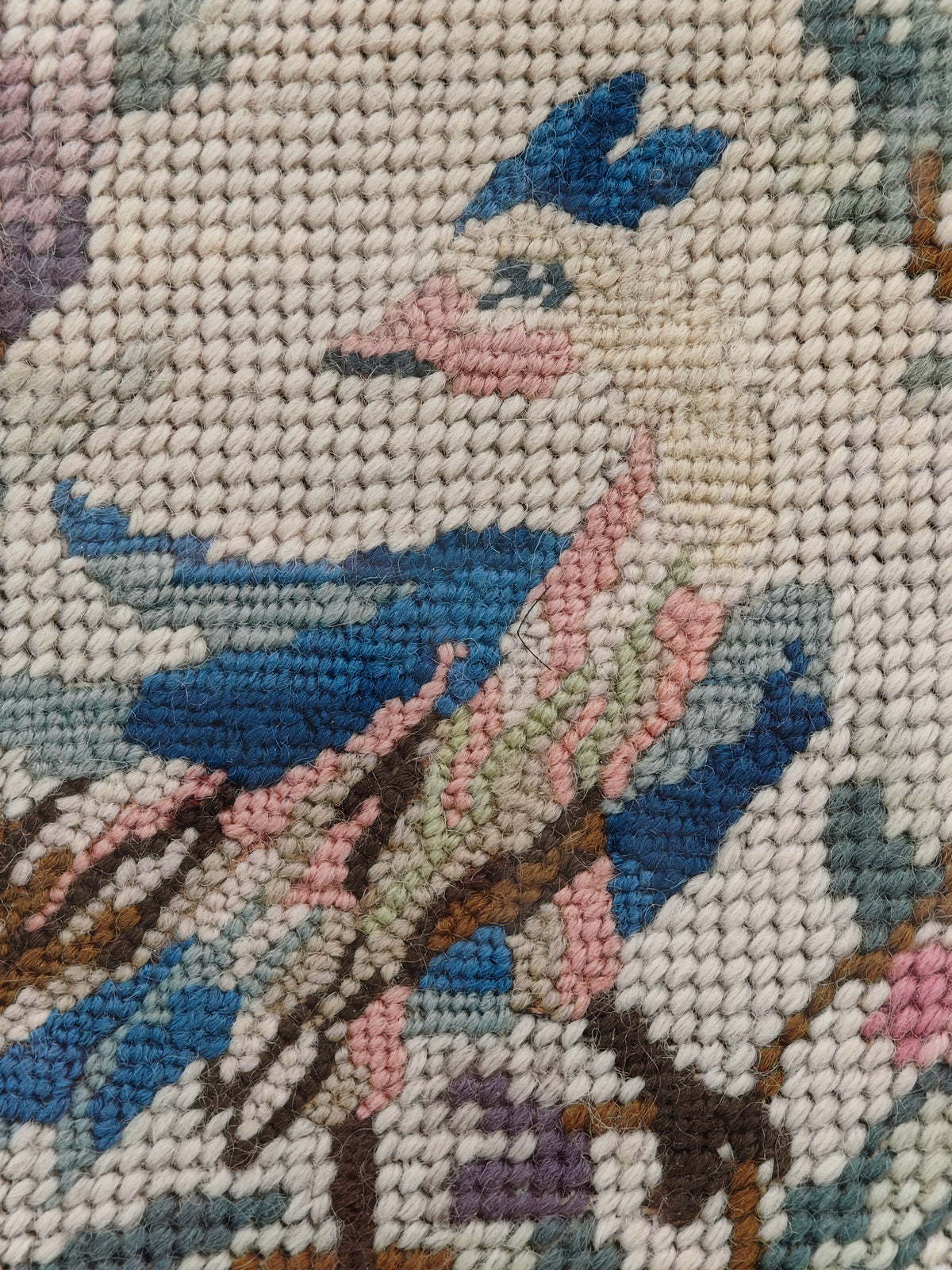 Close-up of a needlepoint canvas featuring a bird design with blue, pink, brown, and cream stitched details.
