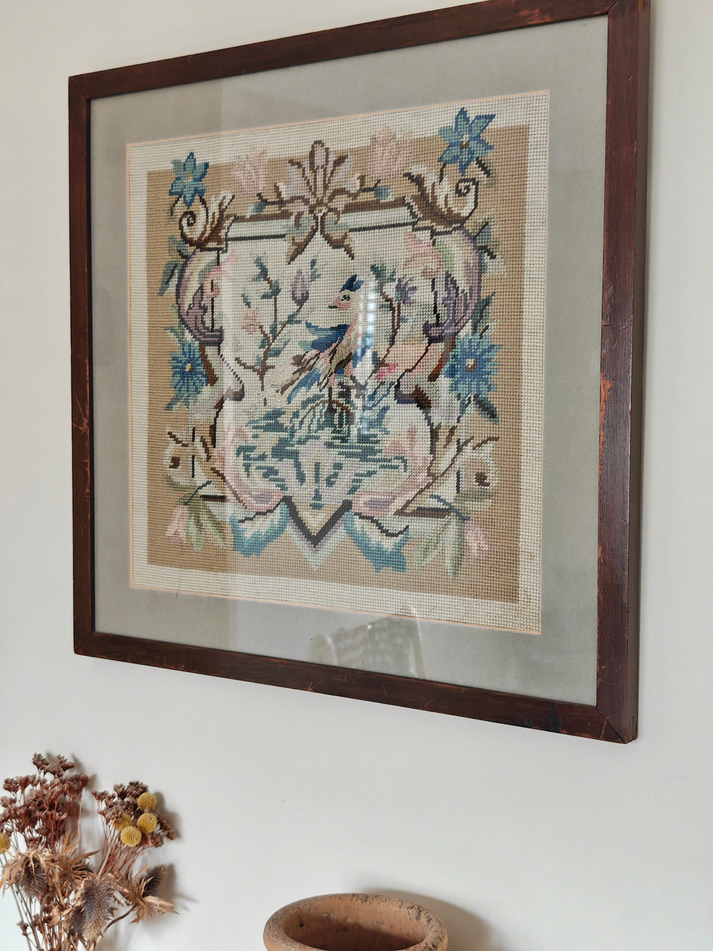 A framed needlepoint artwork featuring a bird and floral scroll design in muted blue, pink, and beige tones.