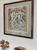 A framed needlepoint artwork featuring a bird and floral scroll design in muted blue, pink, and beige tones.