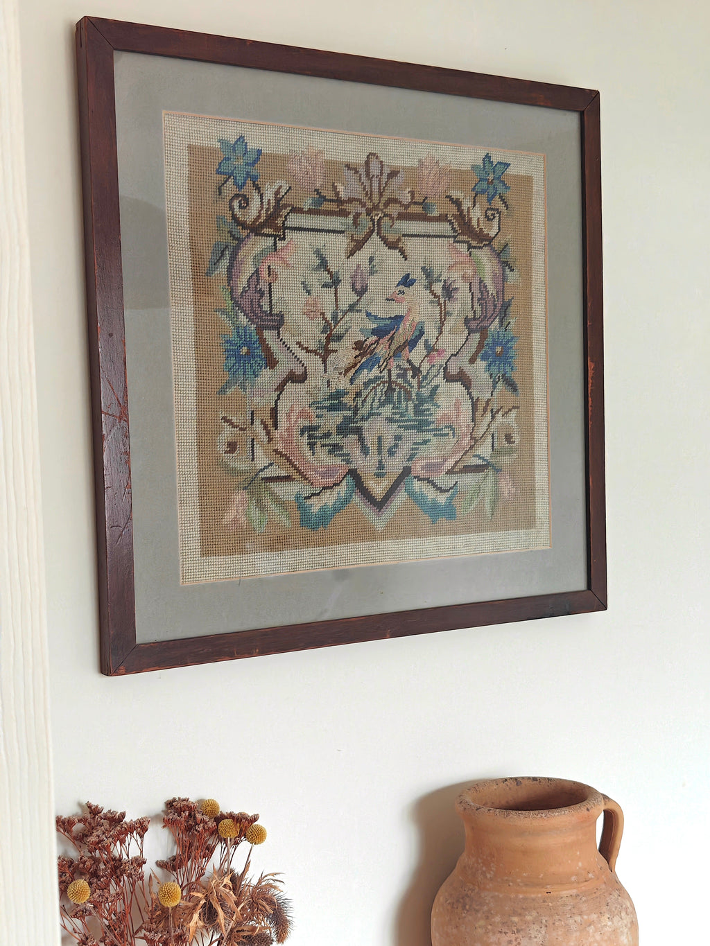 A framed needlepoint artwork featuring a bird and floral scroll design in muted blue, pink, and beige tones.