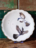 A white plate with scalloped edge and gold trim decorated with two birds in flight with grey, brown and white feathers.