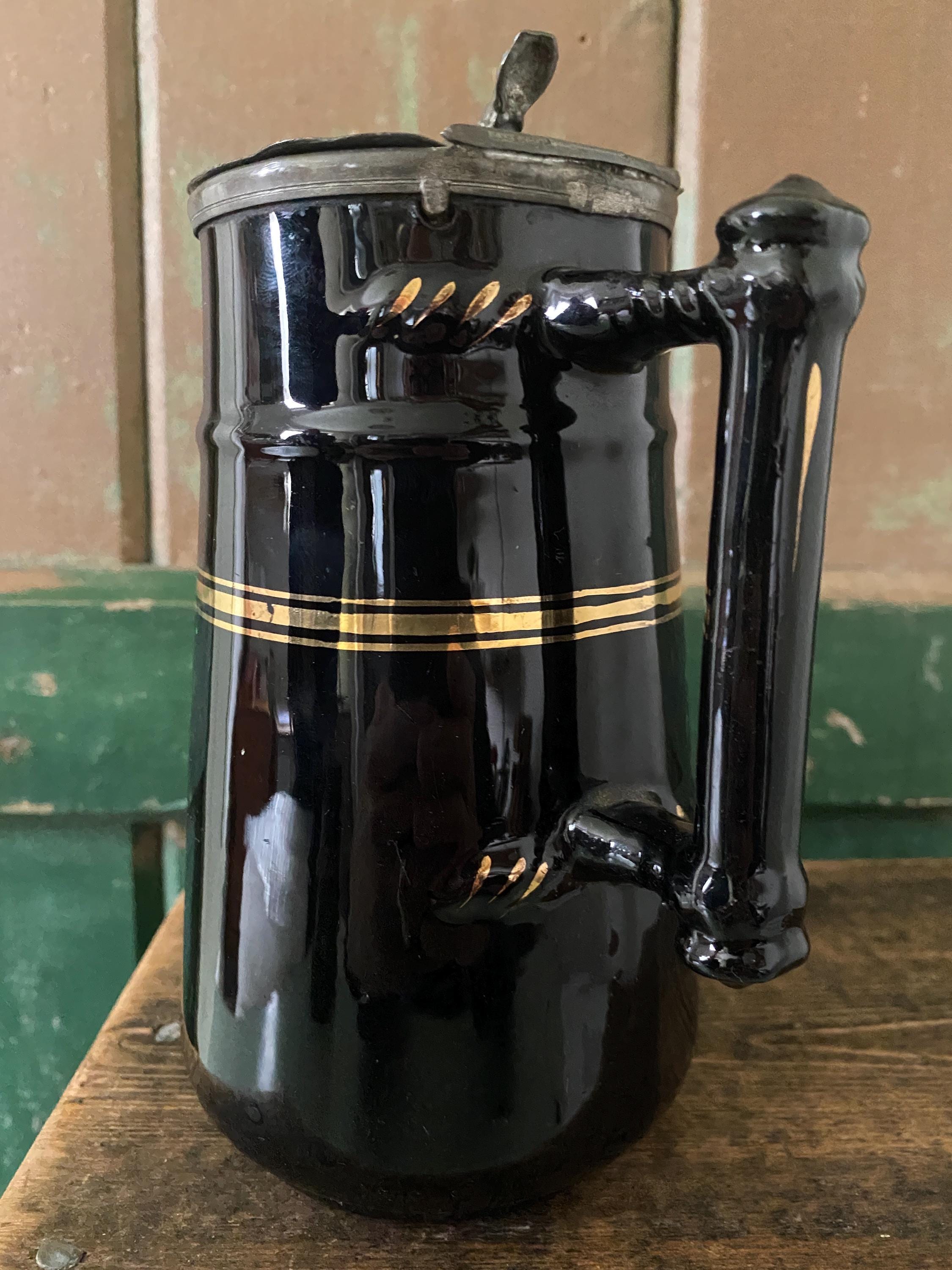 Handle detail of a black ceramic pitcher with gold stripe and hinged metal lid